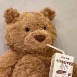 Caramel brown curly fur teddy bear plush toy with wide smile and bead eyes, holding a lychee peach milk carton, cosy kawaii cuddly toy UK